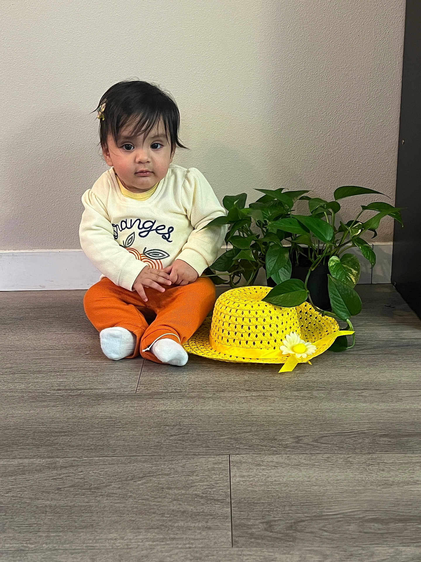 Dhriti joined the competition — help win amazing prizes! baby, child, plant, yellow_hat, floor, indoor, orange_pants, long_sleeve_shirt, curly_hair, leafy_plant, white_socks, flower, hat, sitting, looking_at_camera, neutral_expression, wall, home, casual_clothing, cute