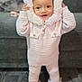 baby, child, pink_clothing, knitted_outfit, hood, smiling, standing, indoor, rug, cozy, cute, happy, person, furniture, home, soft_texture, legs, feet, buttons, curly_hair