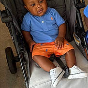 Karter joined the competition — help win amazing prizes! toddler, child, stroller, blue_shirt, orange_shorts, white_sneakers, indoor, carpet, seated, curious, baby, clothing, footwear, person, portrait, casual, baby_fashion, wheel, seat, young_child