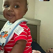 Karter is registered to the contest to win money with this photo: baby, child, video_call, smiling, indoor, bed, blanket, clothing, person, face, infant, technology, screen, digital, communication, happy, portrait, red_shirt, striped_clothing, furniture