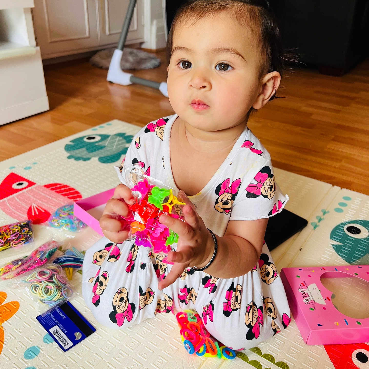Inayat joined the competition — help win amazing prizes! bracelet, child, colorful, craft_supplies, dress, expression, floor, furniture, hair_tie, indoor, minnie_mouse, person, plastic_clips, play_mat, room, sitting, toddler, toy, wooden_floor, young_child