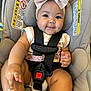baby, child, car_seat, safety_harness, pink_bow, smile, chubby_cheeks, infant, cute, clothing, footwear, seatbelt, straps, indoor, person, skin, face, hand, leg, closeup