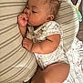 baby, sleeping, thumb_sucking, curly_hair, pillow, striped, floral, bedspread, infant, cute, peaceful, child, indoors, resting, soft_lighting, cozy, clothing, onesie, earring, skin