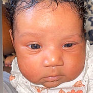 Kamora is registered to the contest to win money with this photo: baby, infant, face, curly_hair, eyes, skin, clothing, floral_pattern, white_outfit, cute, portrait, closeup, person, child, newborn, expressionless, indoors, leopard_print, background, soft_lighting