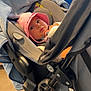 baby, pink_hat, stroller, blanket, indoor, child, infant, face, wide_eyes, seat, black_frame, wheel, cozy, curious, window, safety_sign, floor, clothing, person, portrait