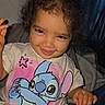 blue_character, cartoon, child, couch, curly_hair, curtain, dark_hair, eyes, face, grinning, home, indoor, photography, pink_clothing, portrait, sitting, smiling, stitch, t_shirt, toddler