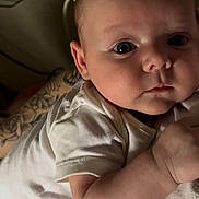 Riley is registered to the contest to win money with this photo: baby, infant, child, person, face, skin, arm, tattoo, onesie, clothing, lying_down, indoor, closeup, curious, expression, human, newborn, cute, young, portrait