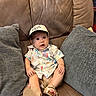 baby, child, infant, shirt, cap, hat, couch, sofa, cushion, brown, gray, footwear, sandals, indoor, furniture, person, cute, sitting, expression, comfortable