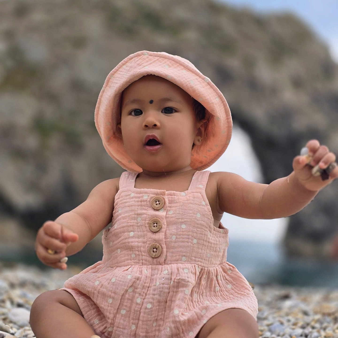 Luniva is registered to the contest to win money with this photo: baby, barefoot, child, closeup, curious, cute, hat, infant, nature, outdoor, pebble_beach, polka_dot, portrait, rock_arch, romper, sea, sitting, summer, sun_hat, water