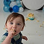 toddler, child, cake, blue_eyes, messy, birthday, balloons, celebration, snack, denim_overalls, curly_hair, indoor, happy, cute, portrait, face, hand, food, smile, party