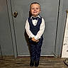 child, boy, smile, suit, bow_tie, formal_wear, standing, door, indoor, floor, wood_floor, happy, portrait, person, clothing, shoes, young, cute, fashion, pose
