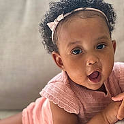 Talitha joined the competition — help win amazing prizes! baby, child, curly_hair, headband, pink_clothing, knitted_outfit, couch, indoor, portrait, close_up, expression, soft_lighting, resting, infant, cute, person, skin, eyes, mouth_open, comfortable