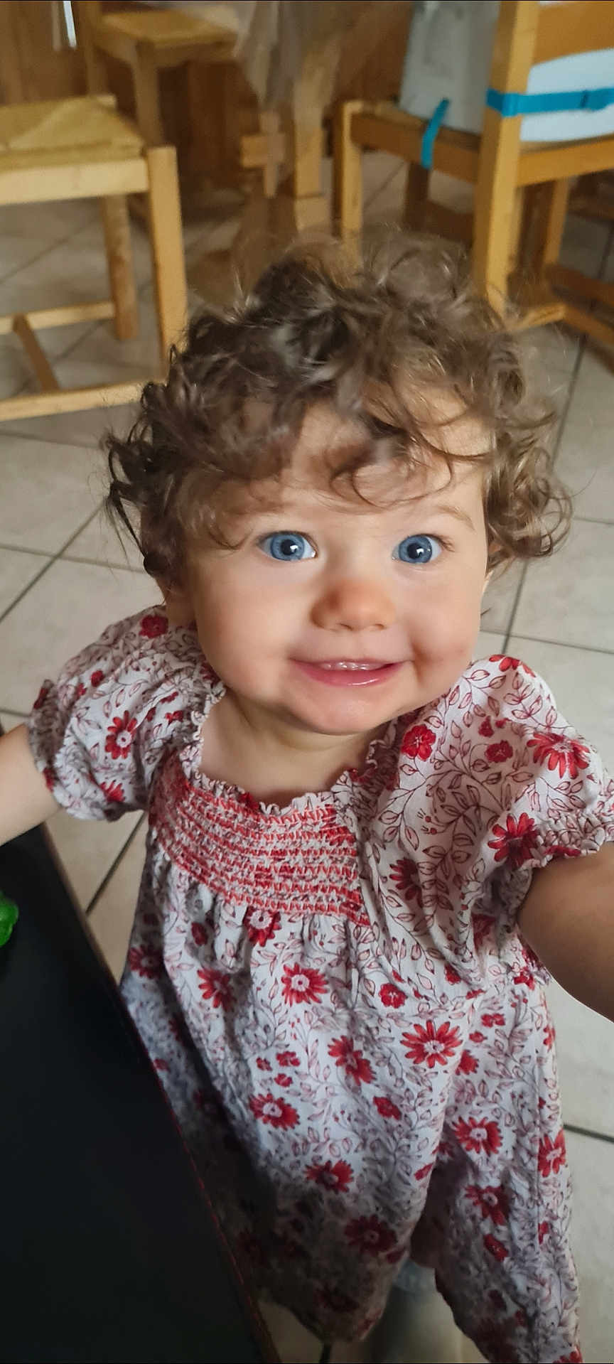 Mailey participe au concours pour gagner de l'argent avec cette photo : child, toddler, curly_hair, blue_eyes, smile, floral_dress, indoor, tile_floor, wooden_chair, person, cute, happy, portrait, young_child, clothing, baby, face, standing, casual, bright