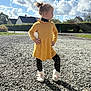 Angela participe au concours pour gagner de l'argent avec cette photo : child, girl, yellow_dress, unicorn_slippers, leggings, gravel, outdoor, sunny, sky, clouds, houses, side_view, pose, hair_bun, playful, shadow, nature, residential, daylight, confident