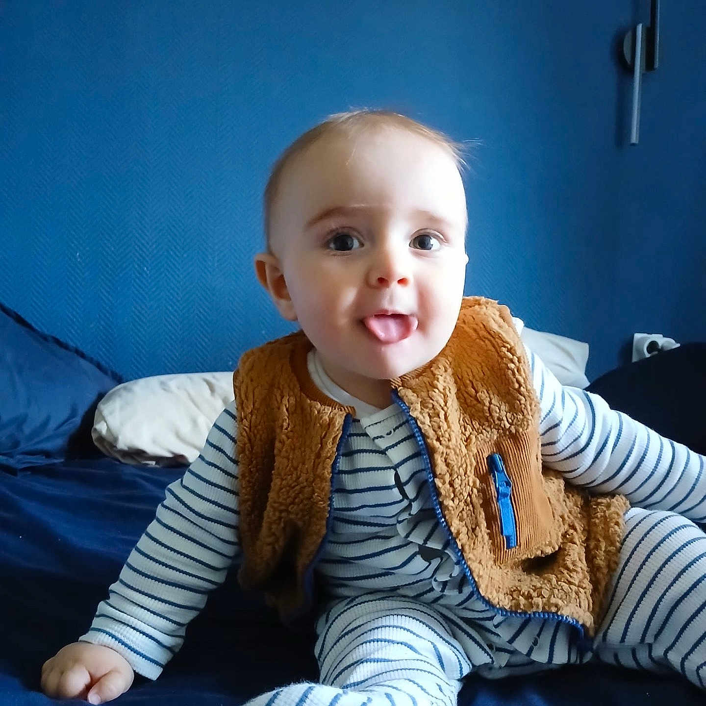 Gabriel participe au concours pour gagner de l'argent avec cette photo : baby, bed, blue_wall, brown_vest, child, cozy, cute, happy, home, indoor, infant, person, playful, portrait, sitting, smiling, soft_lighting, striped_pajamas, tongue_out, young_child
