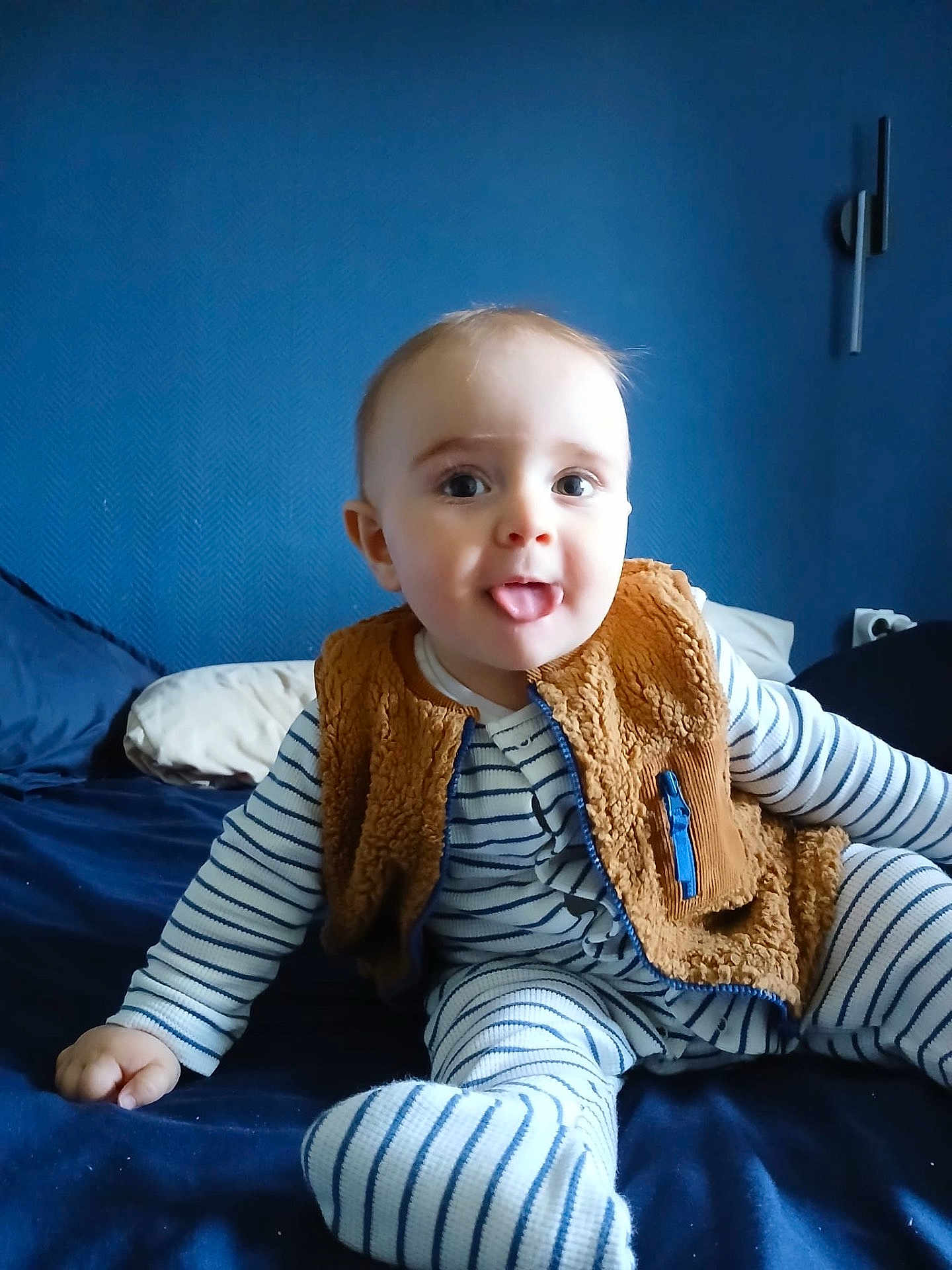 Gabriel participe au concours pour gagner de l'argent avec cette photo : baby, child, striped_pajamas, brown_vest, bed, blue_wall, playful, tongue_out, sitting, indoor, cute, person, infant, smiling, soft_lighting, home, portrait, cozy, happy, young_child