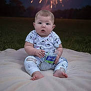 Angel is registered to the contest to win money with this photo: baby, child, infant, sitting, portrait, fireworks, night, outdoor, grass, blanket, water_bottle, drinking, clothing, patterned_shirt, pants, bare_feet, face, surprised_expression, celebration, photography