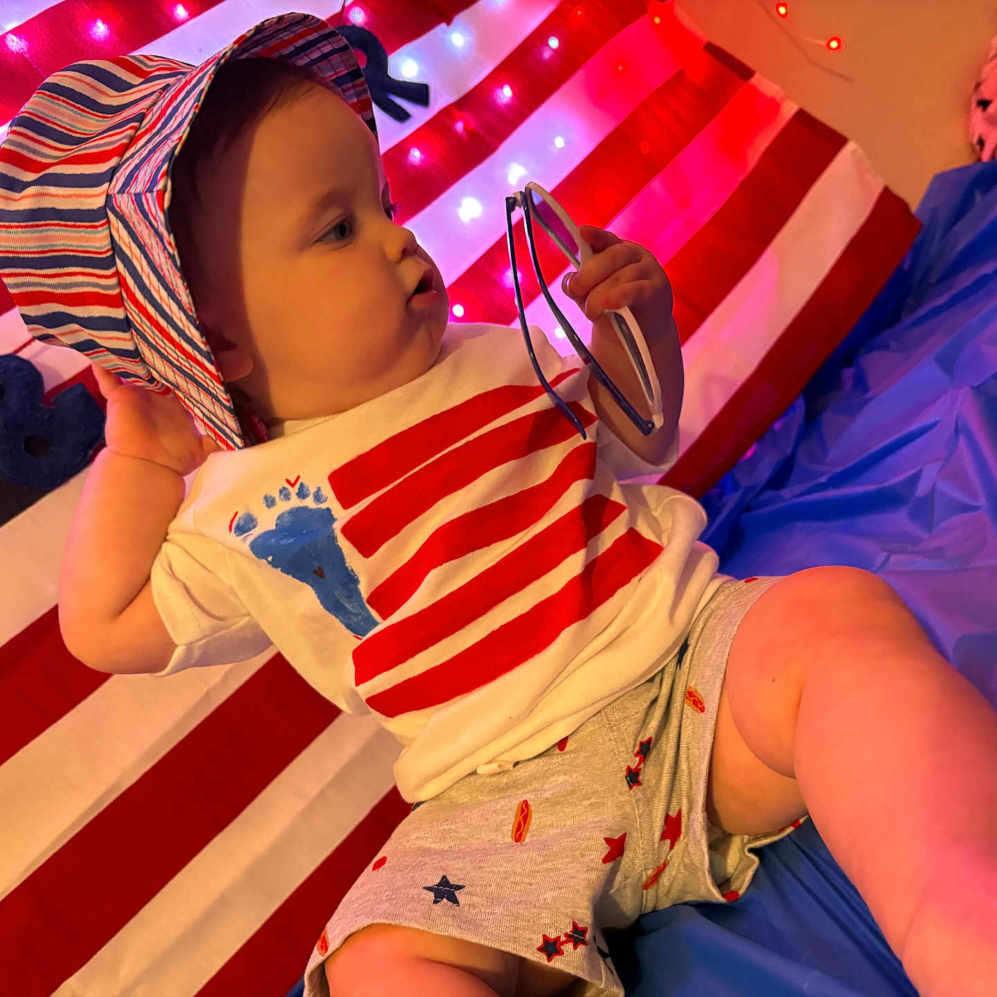 Beckette is registered to the contest to win money with this photo: american_flag, baby, blue, child, curious, cute, fairy_lights, glasses, hat, indoor, infant, patriotic, person, playful, red, shorts, sitting, striped, tshirt, white