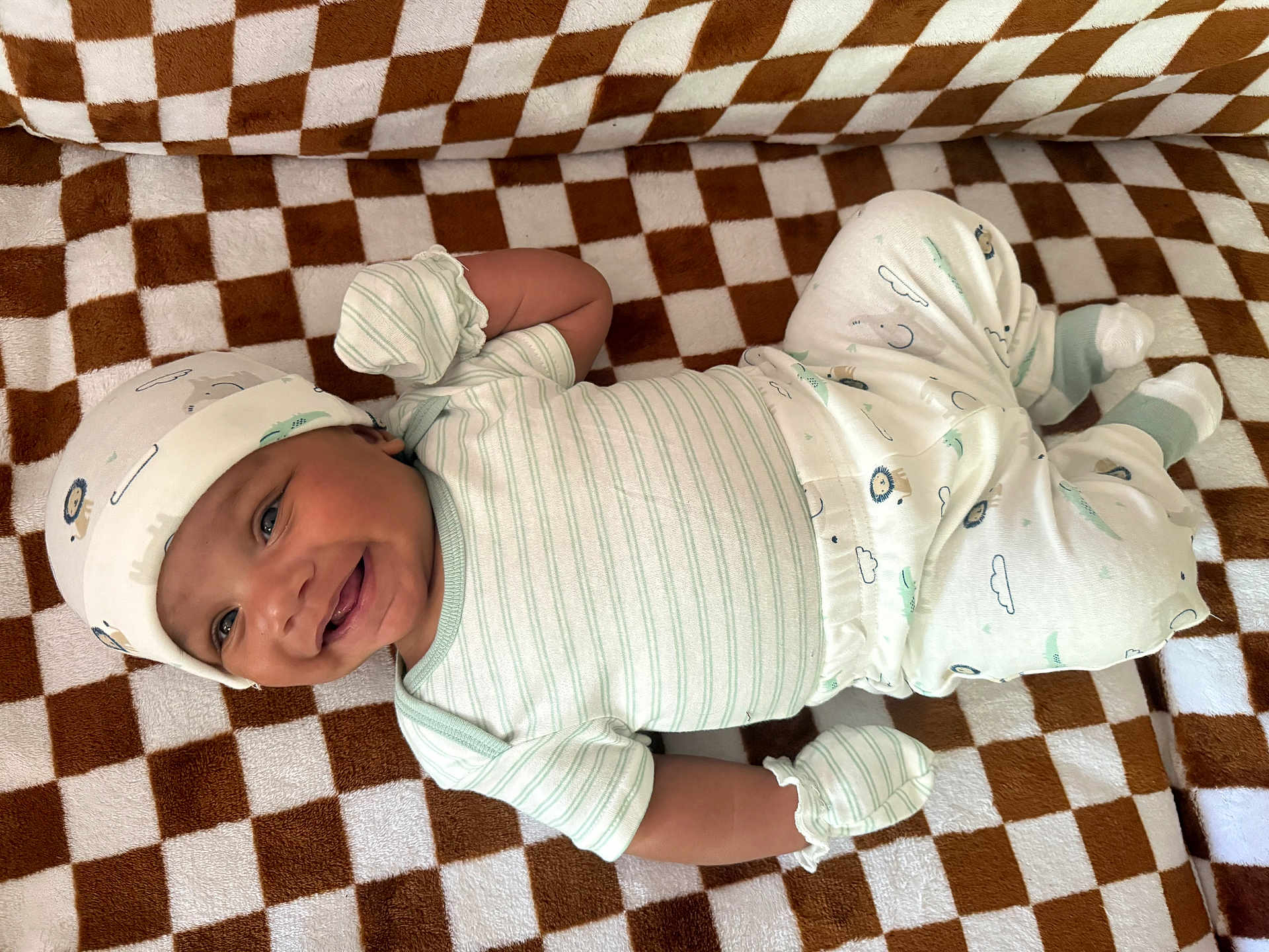 Isaiah is registered to the contest to win money with this photo: baby, smiling, infant, pajamas, hat, mittens, blanket, checkered_pattern, lying_down, happy, cute, child, indoors, face, skin, clothing, newborn, person, comfort, soft