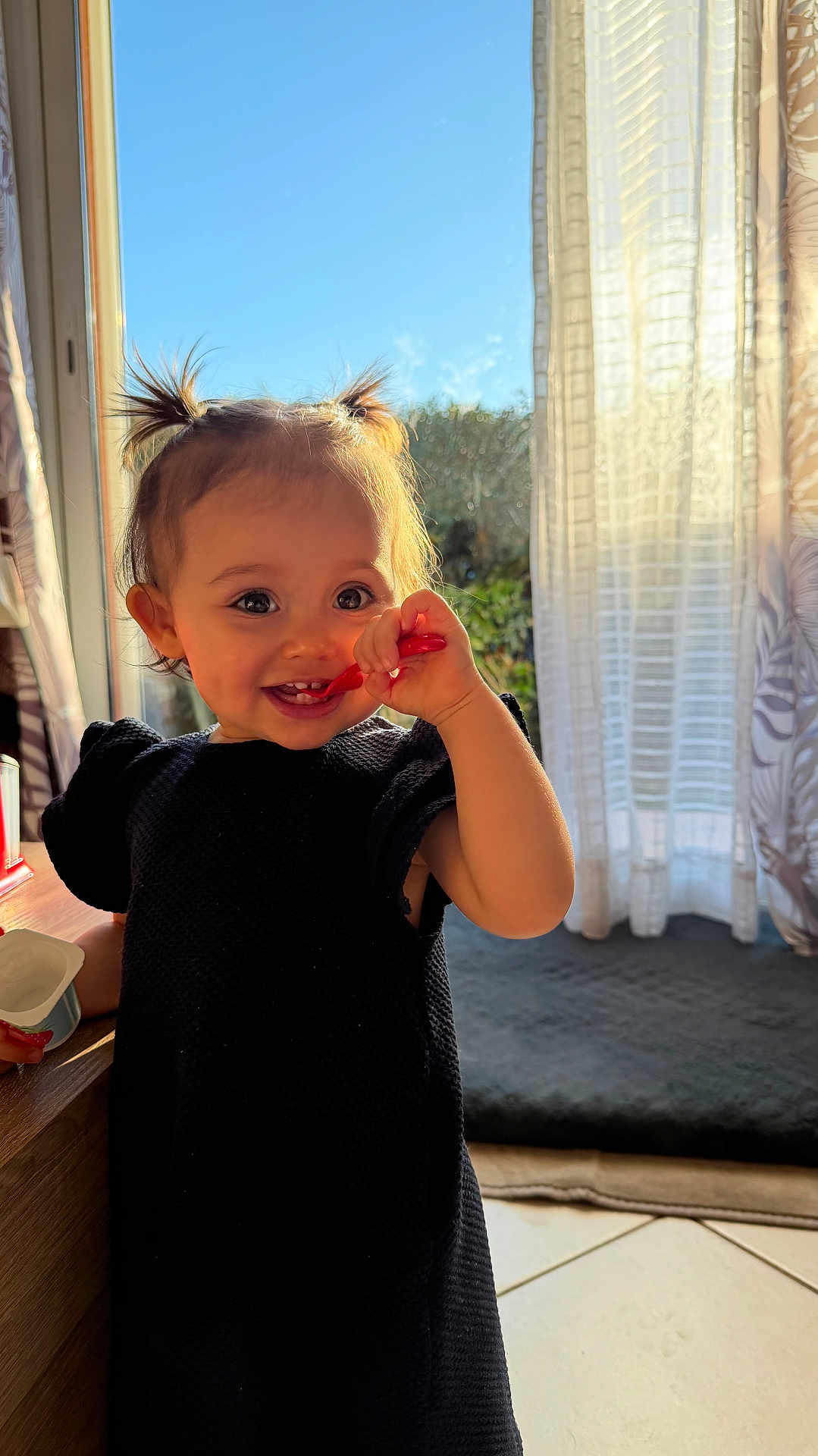 Ghjulia a rejoint le concours — aidez-le/la à gagner de superbes lots ! toddler, child, indoor, smiling, pigtails, yogurt, spoon, black_dress, sunlight, window, curtain, happy, face, person, floor, table, home, daylight, cute, food