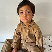 Simon-Léonard a rejoint le concours — aidez-le/la à gagner de superbes lots ! child, boy, sitting, indoors, bed, bedspread, star_pattern, brown_clothing, corduroy, socks, white_socks, green_heart, casual_wear, portrait, young_child, curious_eyes, cross_legged, neutral_expression, plain_wall, cozy