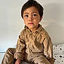 child, boy, sitting, indoors, bed, bedspread, star_pattern, brown_clothing, corduroy, socks, white_socks, green_heart, casual_wear, portrait, young_child, curious_eyes, cross_legged, neutral_expression, plain_wall, cozy
