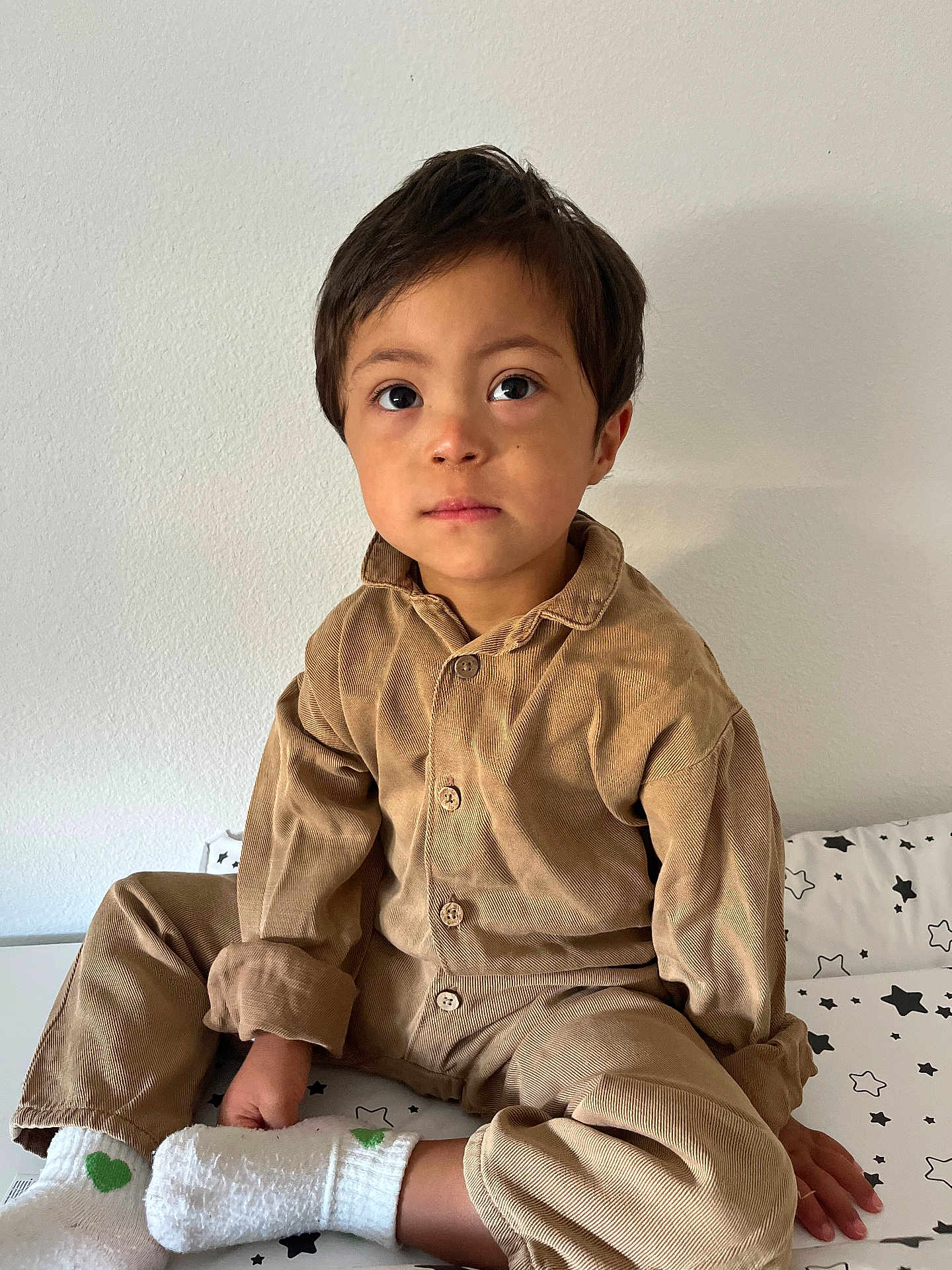 Simon-Léonard a rejoint le concours — aidez-le/la à gagner de superbes lots ! child, boy, sitting, indoors, bed, bedspread, star_pattern, brown_clothing, corduroy, socks, white_socks, green_heart, casual_wear, portrait, young_child, curious_eyes, cross_legged, neutral_expression, plain_wall, cozy