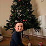 child, toddler, christmas_tree, holiday, decorations, indoors, carpet, smiling, socks, yellow_pants, curly_hair, festive, home, floor, happy, cute, playful, seasonal, celebration, young_child