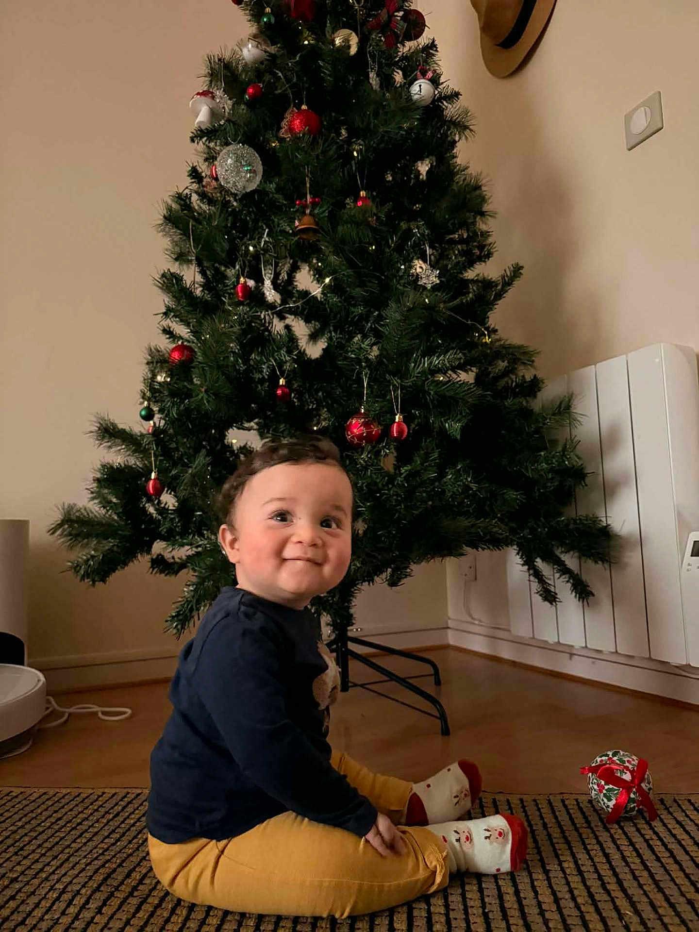 Jad a rejoint le concours — aidez-le/la à gagner de superbes lots ! child, toddler, christmas_tree, holiday, decorations, indoors, carpet, smiling, socks, yellow_pants, curly_hair, festive, home, floor, happy, cute, playful, seasonal, celebration, young_child