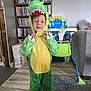 child, dinosaur_costume, green, costume, barefoot, rug, high_chair, monster_design, bookshelf, dvds, living_room, floor, carpet, smile, squinting, person, indoors, furniture, clothing, toy