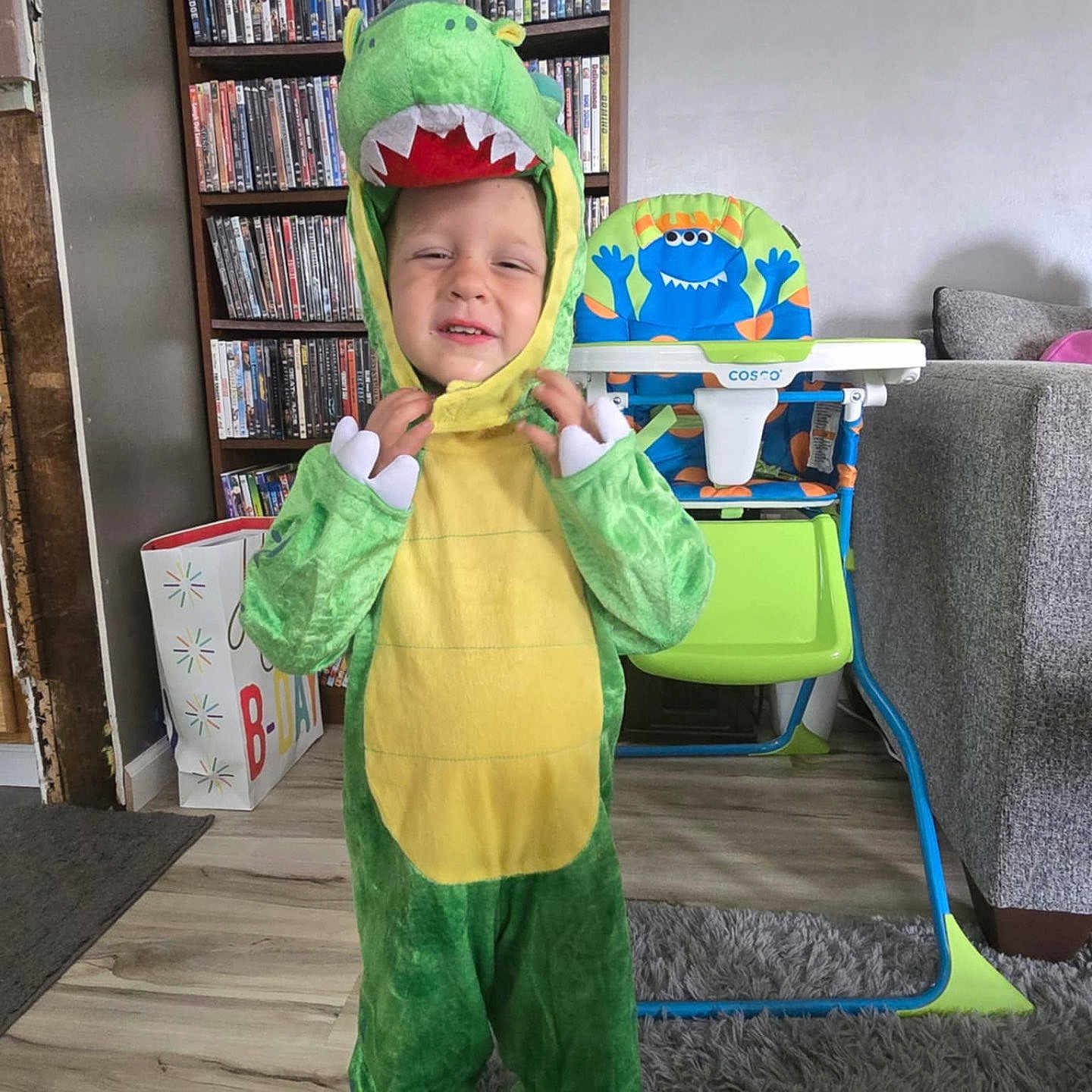 Colby joined the competition — help win amazing prizes! barefoot, bookshelf, carpet, child, clothing, costume, dinosaur_costume, dvds, floor, furniture, green, high_chair, indoors, living_room, monster_design, person, rug, smile, squinting, toy