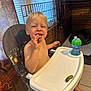 toddler, high_chair, pickle, smiling, indoor, child, blond_hair, sippy_cup, happy, eating, face, furniture, person, footwearless, window_blinds, dog_crate, plastic_tray, wood_paneling, tile_floor, home