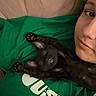Pea is registered to the contest to win money with this photo: animal, black_cat, casual, cat, close_up, companion, cozy, cute, face, feline, green_shirt, indoor, kitten, lying_down, paws, person, pet, relaxed, resting, stretching