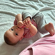 William is registered to the contest to win money with this photo: baby, infant, child, blanket, striped_clothing, gray_pants, sucking_thumb, foot, lying_down, curious, soft_texture, indoor, cute, playful, skin, head, eyes, hand, person, cozy