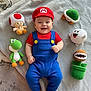 William is registered to the contest to win money with this photo: baby, child, smiling, costume, mario, plush_toys, toy, green_turtle, red_mushroom, green_dinosaur, piranha_plant, ghost, blue_overalls, red_shirt, red_hat, carpet, blanket, indoors, cute, portrait
