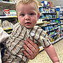 baby, child, person, striped_clothing, hand, store, aisle, shelves, toys, products, shopping, indoor, curious, blonde_hair, blue_eyes, portrait, casual, holding, retail, floor