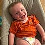 baby, smiling, orange_shirt, diaper, cushion, seat, child, happy, indoor, floor, wooden_floor, toy, infant, cute, person, portrait, skin, face, clothing, relaxing