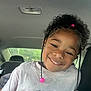 child, smile, curly_hair, beads, tshirt, car_interior, seat, window, natural_light, portrait, happy, person, face, hair_accessory, casual_clothing, young, cute, indoors, seatbelt, head