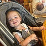 toddler, stroller, smiling, child, baby, person, indoor, restaurant, table, chair, family, casual, clothing, holding, wooden_furniture, seat_belt, happy, face, head, parent