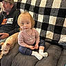 toddler, child, man, dog, couch, blanket, indoor, plush, casual_clothing, cap, hoodie, pet, seated, relaxed, curious, person, home, living_room, white_socks, checkered_pattern