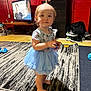 Olivia is registered to the contest to win money with this photo: toddler, child, blue_tutu, barefoot, indoor, rug, toy, dresser, television, red_wall, shoes, carpet, person, furniture, floor, hair_tie, room, clothing, standing, young_child