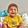 baby, child, smiling, cap, yellow_shirt, fries_graphic, sitting, bed, blanket, blue_package, happy, portrait, indoors, cute, infant, skin, face, hand, foot, clothing