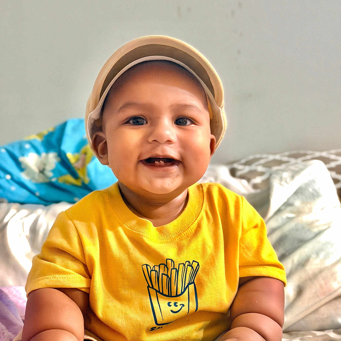 Arhan joined the competition — help win amazing prizes! baby, child, smiling, cap, yellow_shirt, fries_graphic, sitting, bed, blanket, blue_package, happy, portrait, indoors, cute, infant, skin, face, hand, foot, clothing