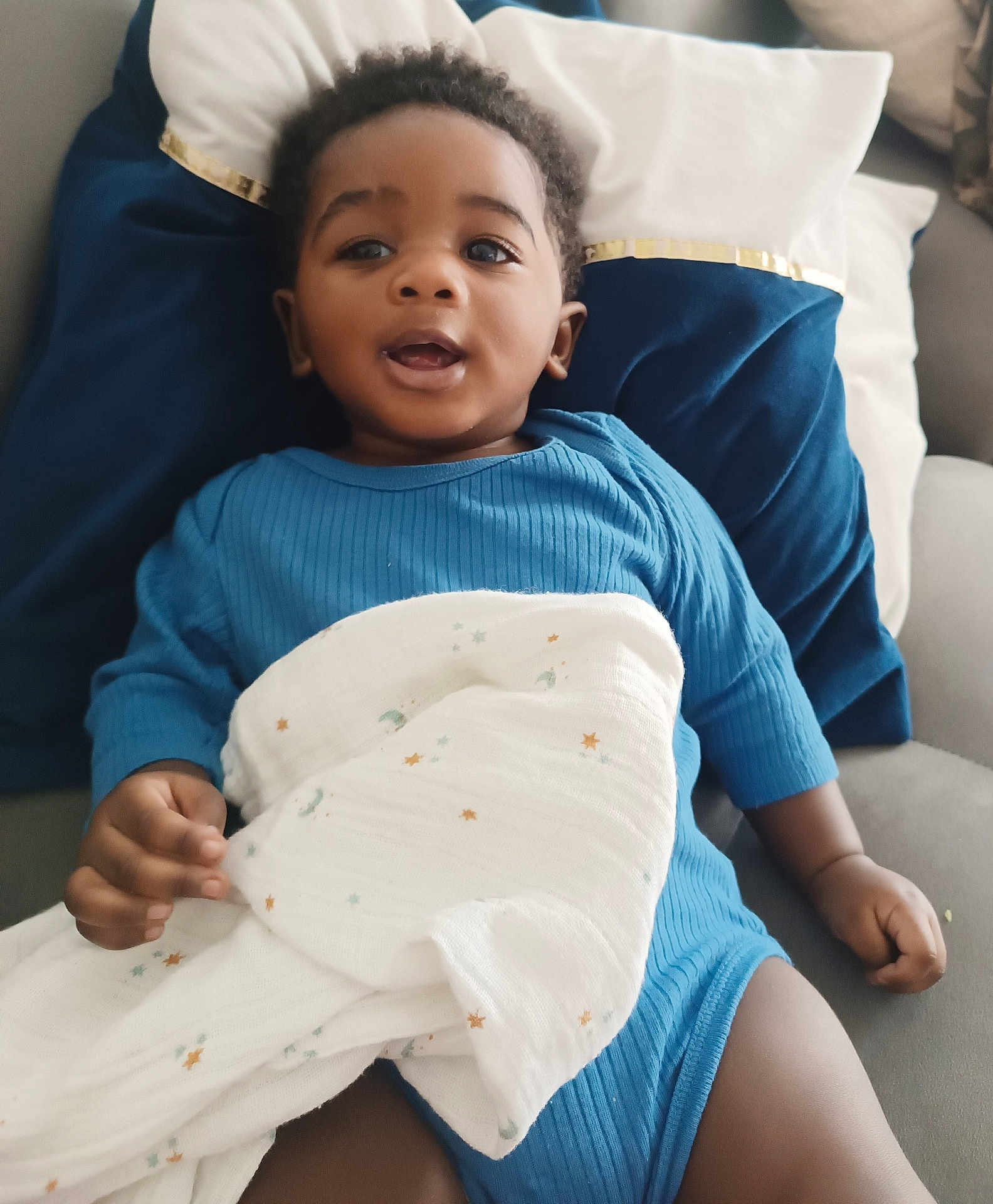 Jaheim a rejoint le concours — aidez-le/la à gagner de superbes lots ! baby, child, blue_clothing, blanket, couch, pillow, indoor, infant, relaxing, smiling, young_child, person, face, hand, cozy, home, soft_fabric, comfort, curious, portrait