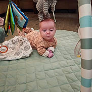 Hazel is registered to the contest to win money with this photo: baby, infant, play_mat, plush_toy, elephant_toy, hanging_toys, floor, wooden_floor, quilted_mat, clothing, person, indoor, cute, child, curious, small_hands, face, head, baby_girl, soft_toys