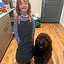 apron, black_apron, cabinet, child, countertop, curly_dog, curly_fur, dog, door, happy, indoor, kitchen, pet, refrigerator, smile, smiling, standing, striped_shirt, wooden_floor, young_child