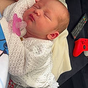 Bella-Leigh is registered to the contest to win money with this photo: newborn, baby, sleeping, pacifier, white_sweater, gray_pants, hand, arm, couch, remote_control, game_controller, red_controller, black_couch, blanket, indoor, person, closeup, resting, cozy, home