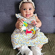 Youvina a rejoint le concours — aidez-le/la à gagner de superbes lots ! baby, child, smiling, toy, colorful_toy, dress, floral_pattern, headband, bow, socks, lace, couch, seated, happy, indoor, person, cute, playful, infant, portrait