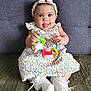 baby, child, smiling, toy, colorful_toy, dress, floral_pattern, headband, bow, socks, lace, couch, seated, happy, indoor, person, cute, playful, infant, portrait