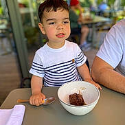 Nino a rejoint le concours — aidez-le/la à gagner de superbes lots ! child, boy, dessert, bowl, spoon, table, striped_shirt, indoor, blurred_background, food, casual, person, sitting, young, cute, meal, cafe, daylight, happy, portrait